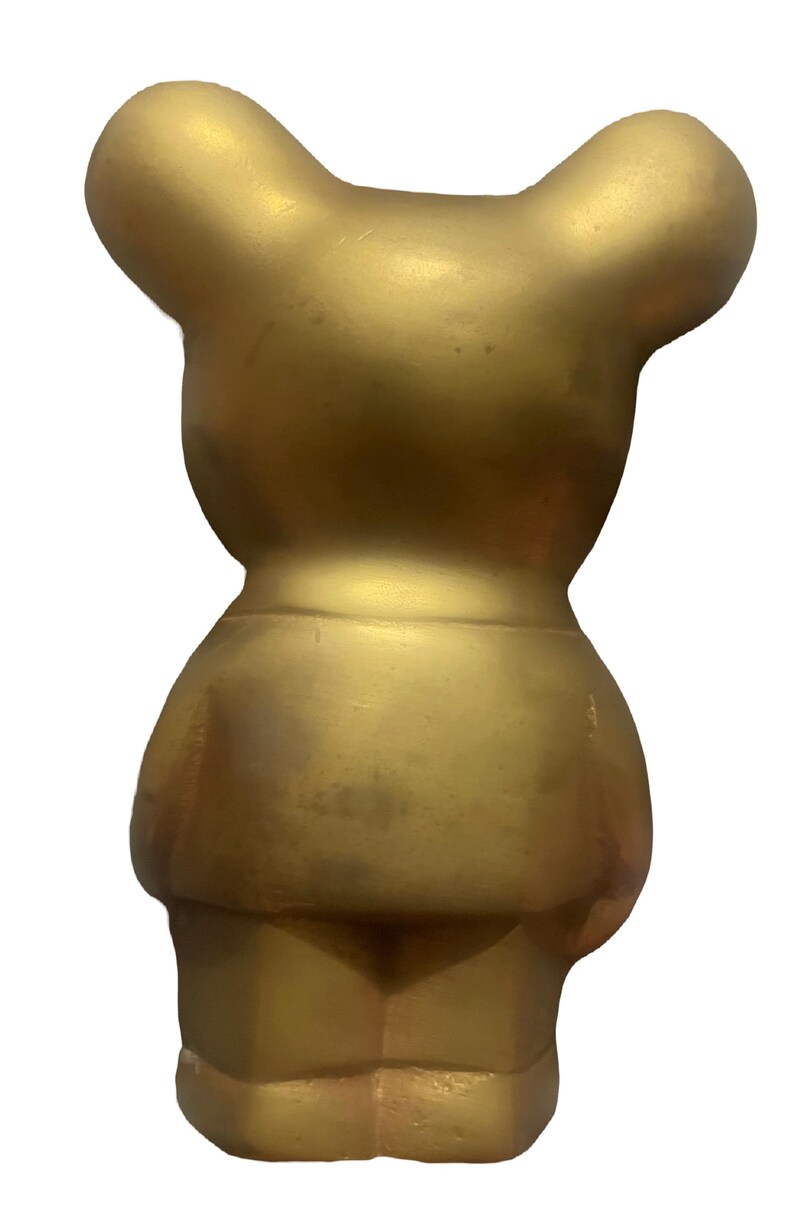 May include: A gold-colored ceramic bear figurine. The bear is standing upright with its arms at its sides. The bear has a simple, rounded design.