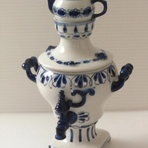 May include: A white and blue ceramic samovar with a small teapot on top. The samovar has a spigot, handles, and is decorated with floral patterns. The teapot and samovar are painted with a dark blue color.