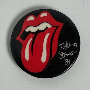 May include: A round black button featuring the iconic red lips and tongue logo of the Rolling Stones. The logo is outlined in white. The words "Rolling Stones '89" are written in white script.