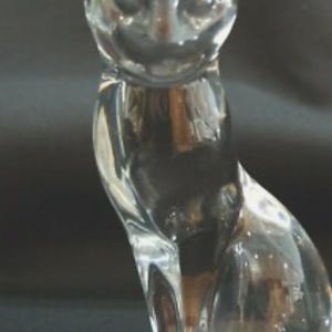 May include: A clear glass cat figurine in a seated position. The cat has pointed ears, a detailed face, and a smooth, reflective surface. The figurine is a decorative item, suitable for display.