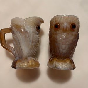 May include: A set of two vintage glass owl figurines. The owl on the left is a pitcher with a brown handle. Both owls have brown eyes and are made of white and brown glass.