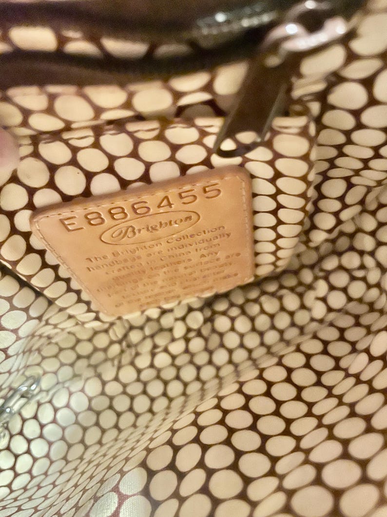 Peut inclure: Gros plan de l'int&eacute;rieur d'un sac &agrave; main brun avec une doublure &agrave; pois blancs et bruns. Une &eacute;tiquette en cuir brun avec le texte "E886455 Brighton The Brighton Collection handbags are individually crafted in China from quality leathers. Any m&eacute;lange on the surface are..." est visible.
