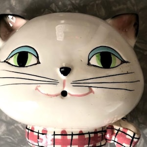 May include: A ceramic cat figurine with a white head, green eyes with blue eyeliner, and black whiskers. The cat has a pink mouth and a red and white checkered collar. The ears are pink inside.