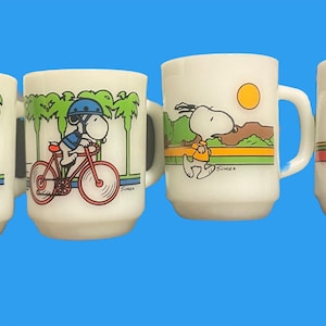May include: Set of four white ceramic mugs with colorful cartoon images of Snoopy. Two mugs feature Snoopy riding a bicycle with palm trees in the background. One mug features Snoopy skateboarding with a sun in the background. The last mug features Snoopy riding a bicycle with a mountain scene in the background.
