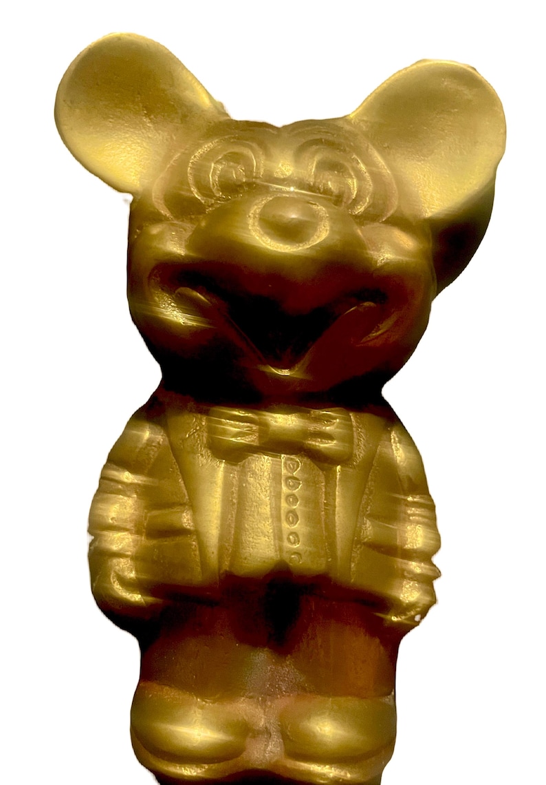 May include: A gold-colored figurine of Mickey Mouse wearing a tuxedo. The figurine is standing with its hands in its pockets and has a big smile on its face.