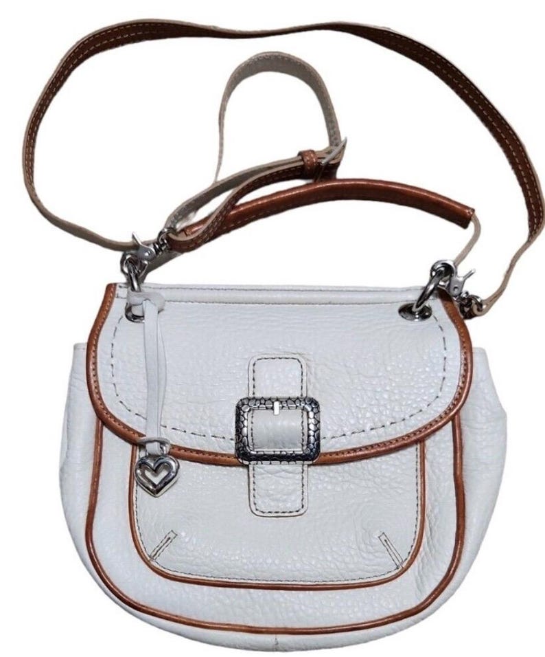 Peut inclure: Sac &agrave; bandouli&egrave;re en cuir blanc avec une bordure en cuir marron et une boucle argent&eacute;e. Le sac est dot&eacute; d'un charm en forme de c&oelig;ur.