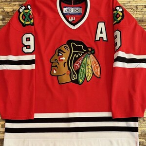May include: Red hockey jersey with black and white stripes. The jersey has the Chicago Blackhawks logo, the number 9, and the letter A on the front. The jersey is made by Reebok.