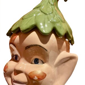 May include: A ceramic cookie jar shaped like an elf's head with a green leaf-shaped lid. The elf has a pink face with blue eyes, a wide smile, and rosy cheeks.