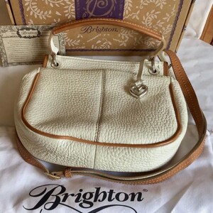 Peut inclure: Un sac &agrave; main Brighton en cuir blanc avec une bandouli&egrave;re en cuir marron et un charm en forme de c&oelig;ur argent&eacute;. Le sac a une poign&eacute;e sup&eacute;rieure et une bandouli&egrave;re. Le sac est pos&eacute; sur une surface en tissu blanc avec le logo Brighton.