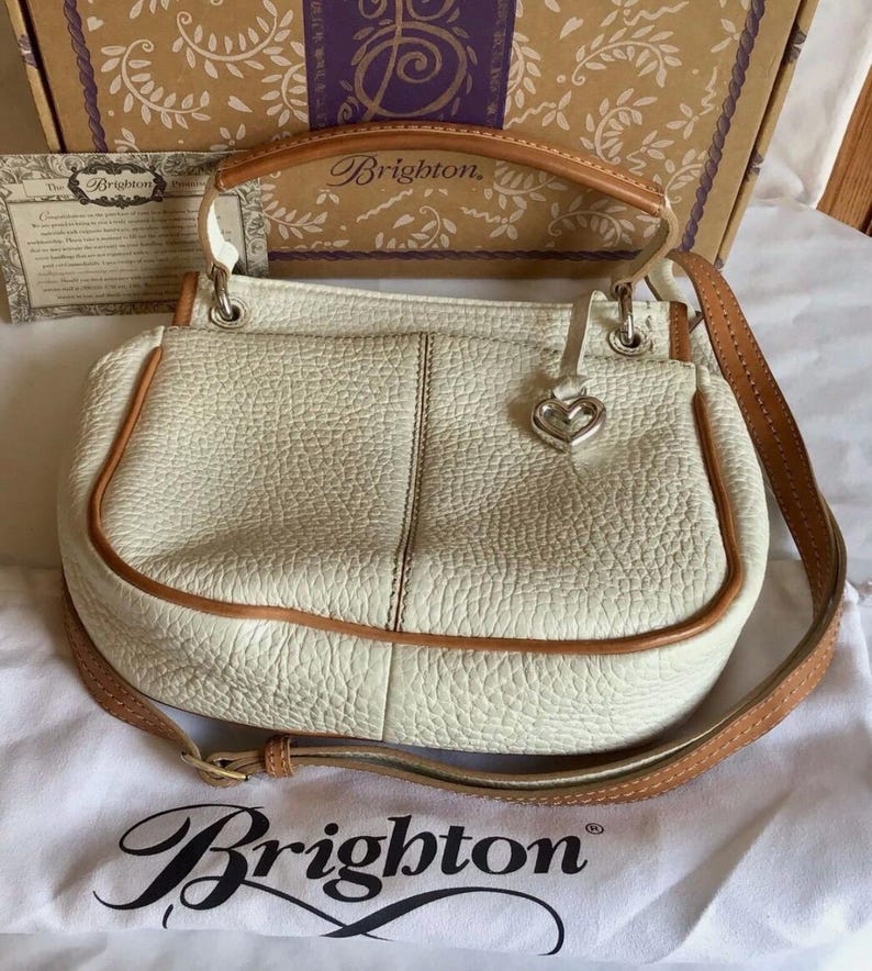 Peut inclure: Un sac &agrave; main Brighton en cuir blanc avec une bandouli&egrave;re en cuir marron et un charm en forme de c&oelig;ur argent&eacute;. Le sac a une poign&eacute;e sup&eacute;rieure et une bandouli&egrave;re. Le sac est pos&eacute; sur une surface en tissu blanc avec le logo Brighton.