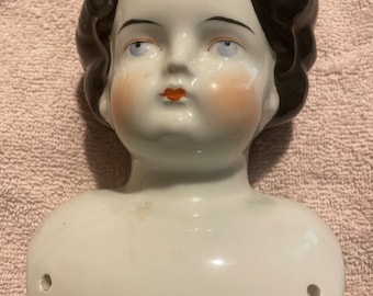 May include: A porcelain doll head with black painted hair and blue eyes. The doll has a painted blush on its cheeks and a red painted mouth. The doll head is attached to a porcelain bust.