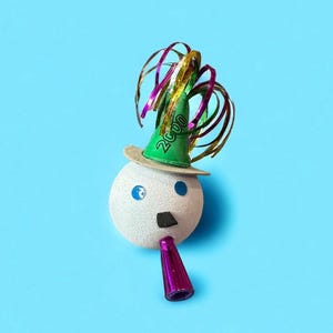 May include: A whimsical, festive decoration resembling a face, set against a bright blue background. It features a white, textured sphere with blue eyes, a black nose, and a purple party horn. A green party hat with "2000" printed on it and metallic streamers complete the design.