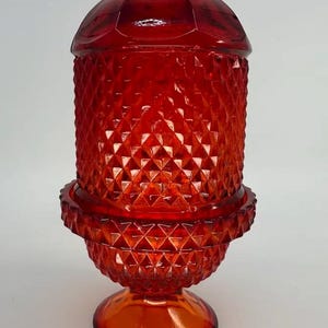 May include: A vintage red glass candle holder with a diamond pattern. The candle holder has a round base, a cylindrical body, and a domed lid. The glass is a deep, rich red color.