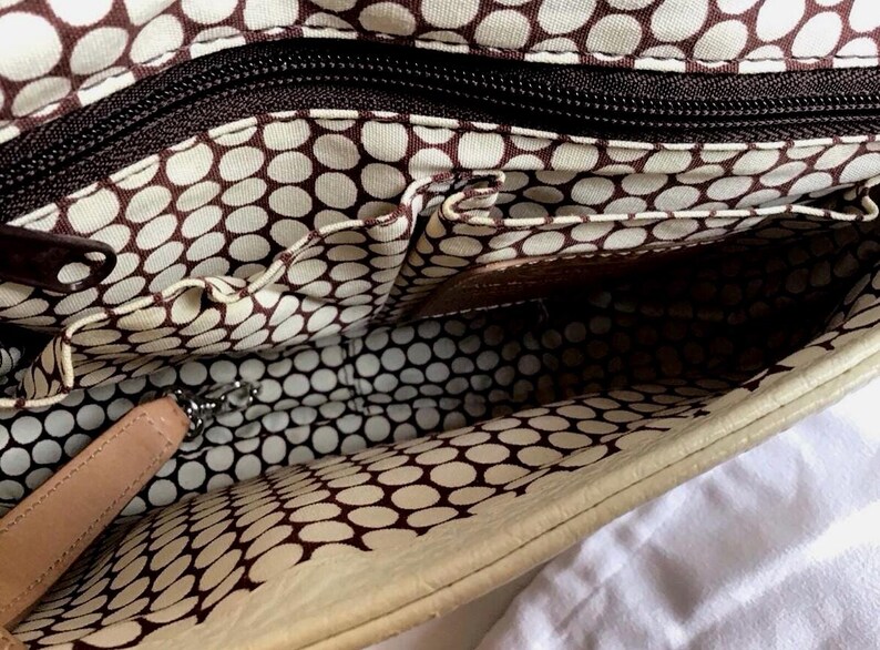 Peut inclure: L'int&eacute;rieur d'un sac &agrave; main en cuir blanc avec une doublure &agrave; pois blancs et bruns. Le sac a une fermeture &agrave; glissi&egrave;re et plusieurs poches.