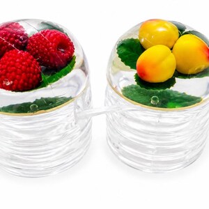 May include: Two clear, ribbed glass containers with dome-shaped lids. One lid features embedded red raspberries and green leaves, while the other contains yellow and orange stone fruits with green leaves. Each container includes a clear glass spoon.