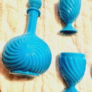 May include: A set of three turquoise blue glass decanters with a swirl design. The decanters have a round body and a tall, slender neck. The set includes a large decanter with a stopper and two smaller decanters.