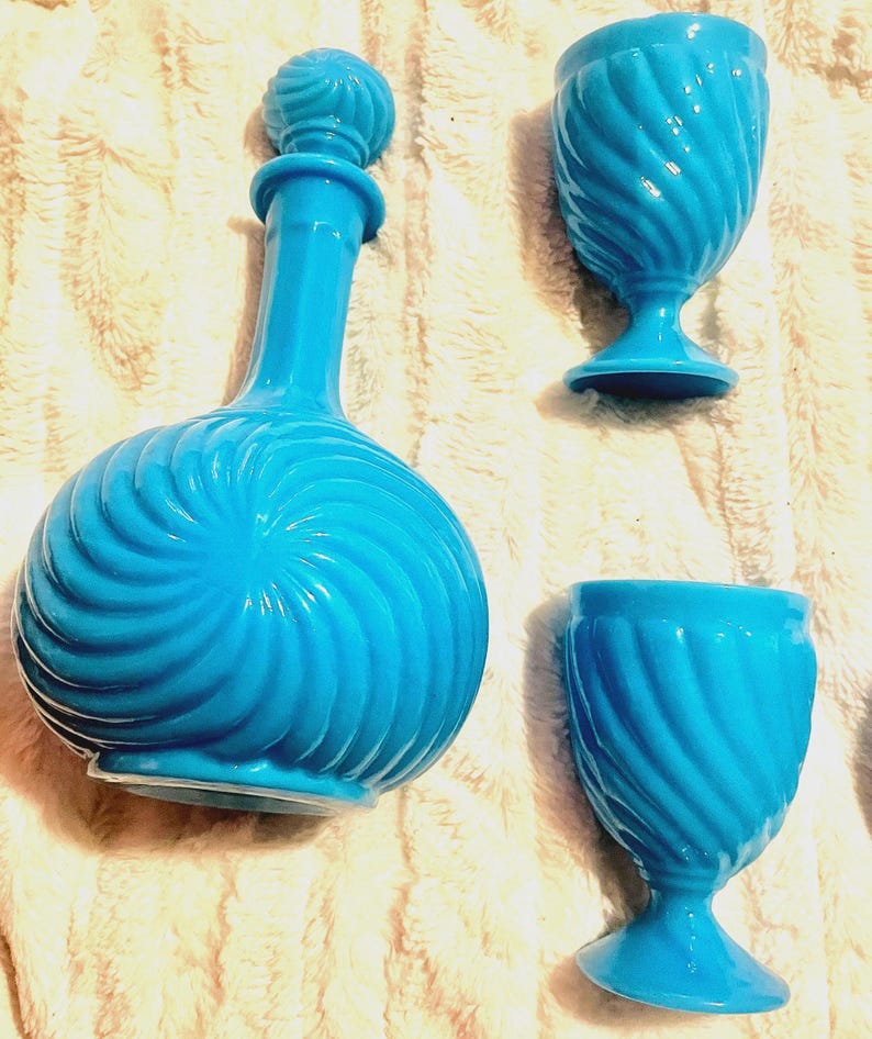 May include: A set of three turquoise blue glass decanters with a swirl design. The decanters have a round body and a tall, slender neck. The set includes a large decanter with a stopper and two smaller decanters.