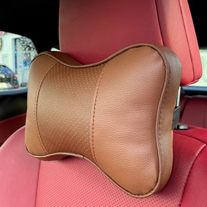 Peut inclure: Appui-t&ecirc;te de voiture marron avec une section centrale perfor&eacute;e et des coutures. L'appui-t&ecirc;te est fix&eacute; &agrave; un si&egrave;ge de voiture rouge. Con&ccedil;u pour le confort et le soutien en voiture.