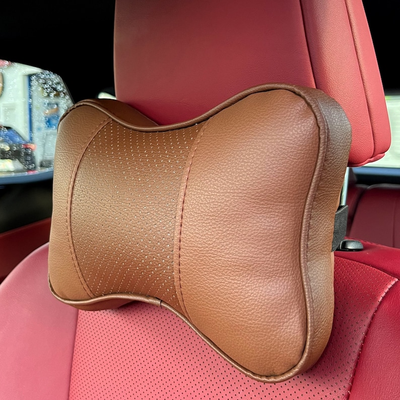 Peut inclure: Appui-t&ecirc;te de voiture marron avec une section centrale perfor&eacute;e et des coutures. L'appui-t&ecirc;te est fix&eacute; &agrave; un si&egrave;ge de voiture rouge. Con&ccedil;u pour le confort et le soutien en voiture.