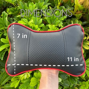 Peut inclure: Coussin de t&ecirc;ti&egrave;re de voiture noir avec garniture rouge, mesurant 28 cm de long et 18 cm de large. Le coussin a une section centrale perfor&eacute;e et le mot "DIMENSIONS" est &eacute;crit en blanc.