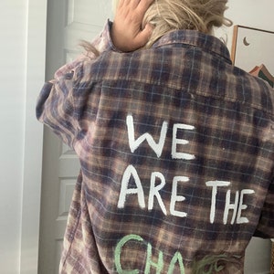 Può includere: Una camicia di flanella a quadri marrone e nero con le parole "We Are The Change" scritte in vernice bianca sul retro.