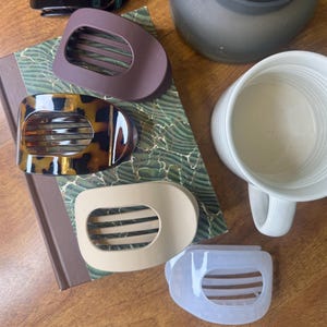 May include: An assortment of hair clips in various colors, including black, brown, tortoiseshell, beige, and light blue. The clips have a unique design with a curved shape and horizontal slits. A candle and a mug are also present.