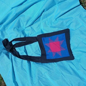 May include: A black fabric tote bag with a blue, pink, and purple star design. The bag has a long strap that can be tied.