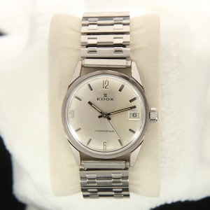 Puede incluir: Un reloj de pulsera plateado con una esfera blanca y agujas negras. El reloj tiene una correa plateada y dice "EDOX HYDROSTAR" en la esfera.