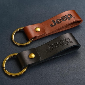 May include: Two leather keychains, one brown and one black, each with the word "Jeep" embossed. The keychains have gold-colored metal rings and rivets. The background is a dark blue textured surface.