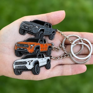 May include: Three miniature truck keychains in gray, orange, and white are connected to a silver key ring. The keychains are detailed with black accents, showcasing the truck's design. The key ring is held against a blurred green background.