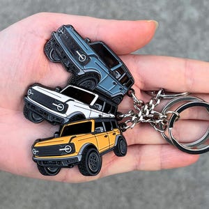 May include: A keychain featuring three miniature SUV car charms in blue, white, and yellow. The charms are attached to a silver chain and key rings. The cars have black tires and detailed designs.