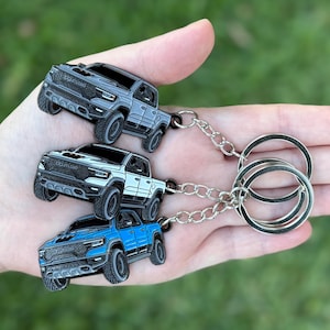 May include: Three metal keychains shaped like pickup trucks in gray, white, and blue. Each keychain has a silver chain and a large silver keyring. The keychains are held in a hand against a blurred green background.