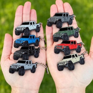Op de afbeelding: Een verzameling miniatuur Jeep truck sleutelhangers in verschillende kleuren, waaronder wit, grijs, blauw, zwart, groen en rood. Elke sleutelhanger heeft een gedetailleerd truckontwerp met zwarte wielen en een zilveren ketting.