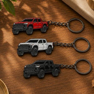 May include: Three metal keychains featuring a pickup truck design, each with a circular ring and chain. The trucks are rendered in red, white, and black enamel finishes, showcasing detailed front grilles, headlights, and rugged tyres on each model.