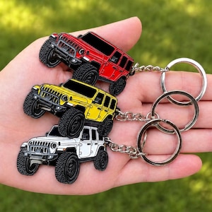 May include: Three Jeep keychains in red, yellow, and white, each with black wheels and a silver keyring. The keychains are held in a hand against a blurred green background.