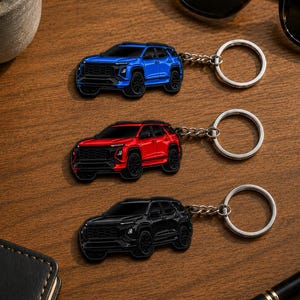 Op de afbeelding: Drie metalen sleutelhangers met een gedetailleerd SUV-ontwerp. Eén sleutelhanger is blauw, één is rood en één is zwart. Elke SUV heeft zwarte wielen en een zwarte grille. De sleutelhangers zijn met korte kettinkjes aan zilverkleurige ringen bevestigd.
