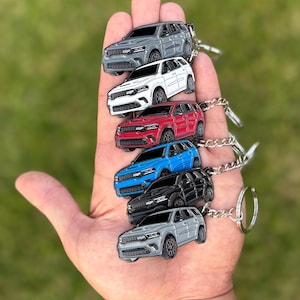 May include: A collection of six SUV keychains in various colors: gray, white, red, blue, black, and silver. Each keychain features a detailed miniature car design with silver keyrings and chains. The keychains are held in a hand against a blurred green background.