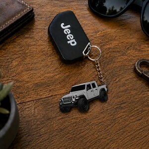 Op de afbeelding: Een zwarte Jeep-sleutelhanger met het woord 'Jeep' in witte letters. Een zilveren Jeep-trucksleutelhanger is bevestigd. De items liggen op een houten oppervlak, met een zonnebril en een portemonnee op de achtergrond.