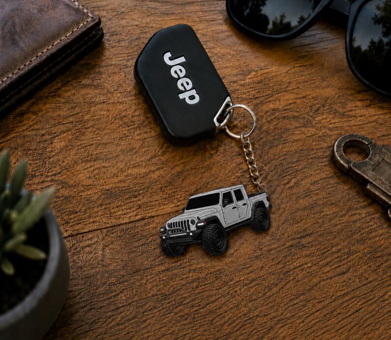 Op de afbeelding: Een zwarte Jeep-sleutelhanger met het woord 'Jeep' in witte letters. Een zilveren Jeep-trucksleutelhanger is bevestigd. De items liggen op een houten oppervlak, met een zonnebril en een portemonnee op de achtergrond.