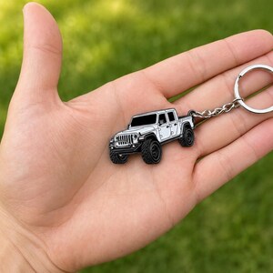 Op de afbeelding: Een wit en zwarte Jeep truck sleutelhanger met een zilveren sleutelring. De sleutelhanger wordt in een hand gehouden tegen een groene achtergrond. De truck heeft zwarte banden en een gedetailleerde grille.