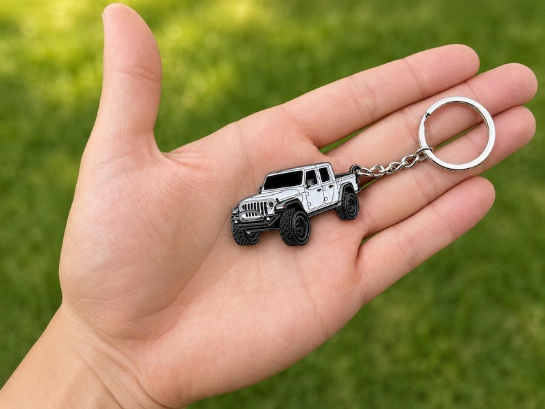 Op de afbeelding: Een wit en zwarte Jeep truck sleutelhanger met een zilveren sleutelring. De sleutelhanger wordt in een hand gehouden tegen een groene achtergrond. De truck heeft zwarte banden en een gedetailleerde grille.