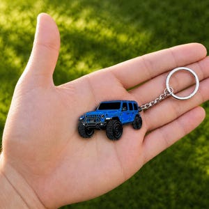 May include: A blue metal keychain shaped like a four-door off-road vehicle with black tires, a front grille, and headlights. The keychain features a silver-toned metal chain and a circular split ring for attaching keys to the vehicle charm.