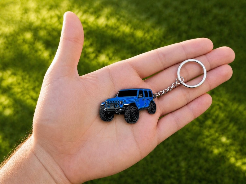 May include: A blue metal keychain shaped like a four-door off-road vehicle with black tires, a front grille, and headlights. The keychain features a silver-toned metal chain and a circular split ring for attaching keys to the vehicle charm.