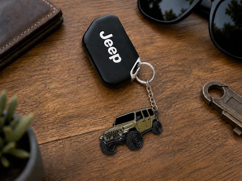 May include: A black Jeep key fob attached to a silver metal keychain featuring a detailed olive green off-road vehicle charm. The charm depicts a four-door SUV with large black tires, a front grille, and tinted windows, connected by a small chain.