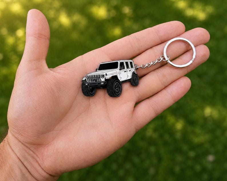 May include: A white off-road vehicle keychain featuring black tires, a black grille, and black windows. The metal charm is attached to a silver-toned circular split ring by a short metal chain, showcasing a detailed miniature SUV design.