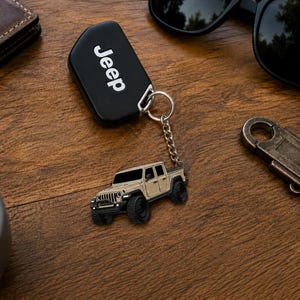 Op de afbeelding: Een zwarte Jeep-sleutelhanger met het woord "Jeep" in witte letters. Een zilveren sleutelhanger is bevestigd aan een klein, beige Jeep-vrachtwagenmodel met zwarte banden. De items liggen op een houten oppervlak.