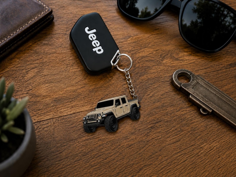 Op de afbeelding: Een zwarte Jeep-sleutelhanger met het woord "Jeep" in witte letters. Een zilveren sleutelhanger is bevestigd aan een klein, beige Jeep-vrachtwagenmodel met zwarte banden. De items liggen op een houten oppervlak.