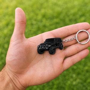 May include: A black metal keychain shaped like a miniature off-road vehicle. The keychain features a detailed, matte black finish with a classic seven-slot grille, round headlights, and rugged tires, attached to a standard silver-toned metal ring.
