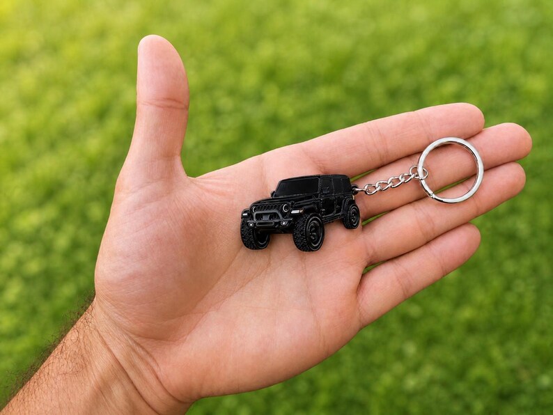 May include: A black metal keychain shaped like a miniature off-road vehicle. The keychain features a detailed, matte black finish with a classic seven-slot grille, round headlights, and rugged tires, attached to a standard silver-toned metal ring.