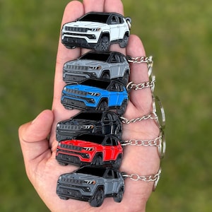 May include: A collection of six Jeep Compass keychains in various colors: white, gray, blue, black, red, and another shade of gray. Each keychain features a detailed miniature car design with a black roof and silver chain.